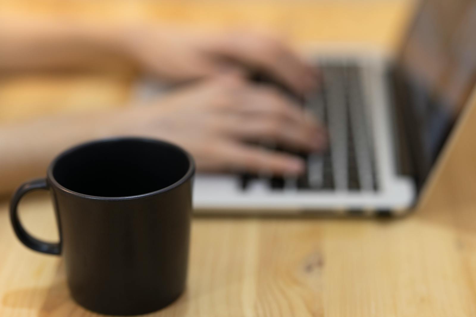Black mug on wooden table with hands typing on laptop, illustrating focused work