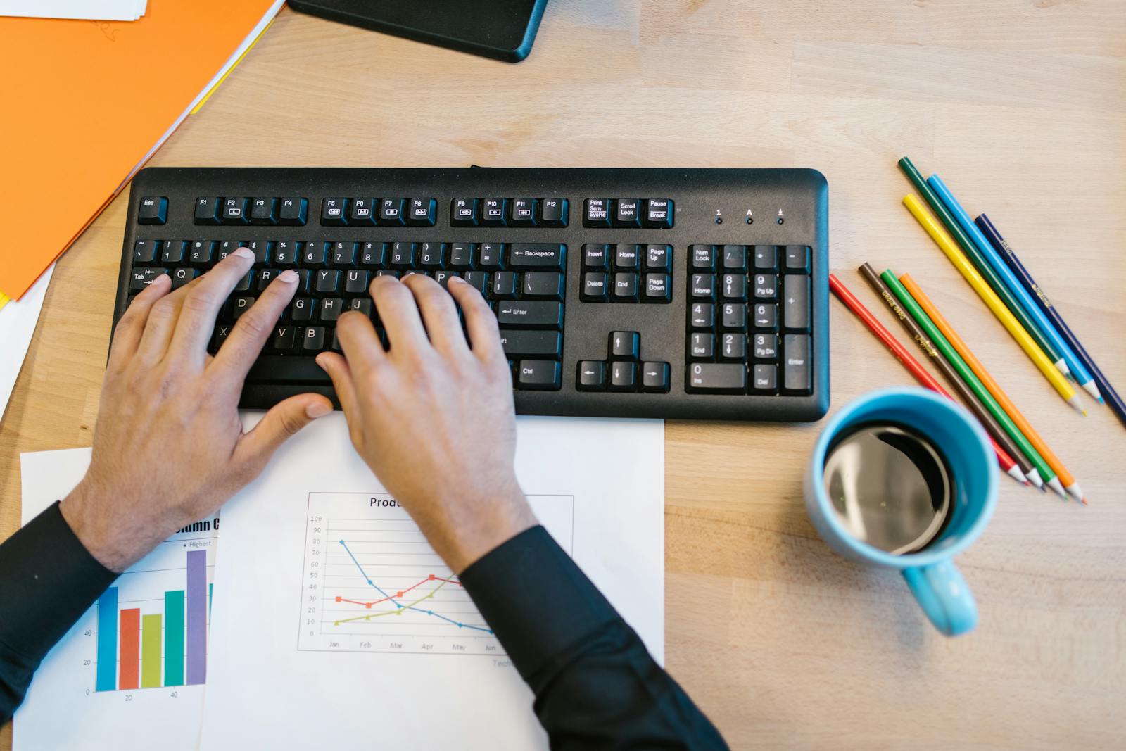 Workspace with hands typing on keyboard, coffee mug, colored pencils, graphs