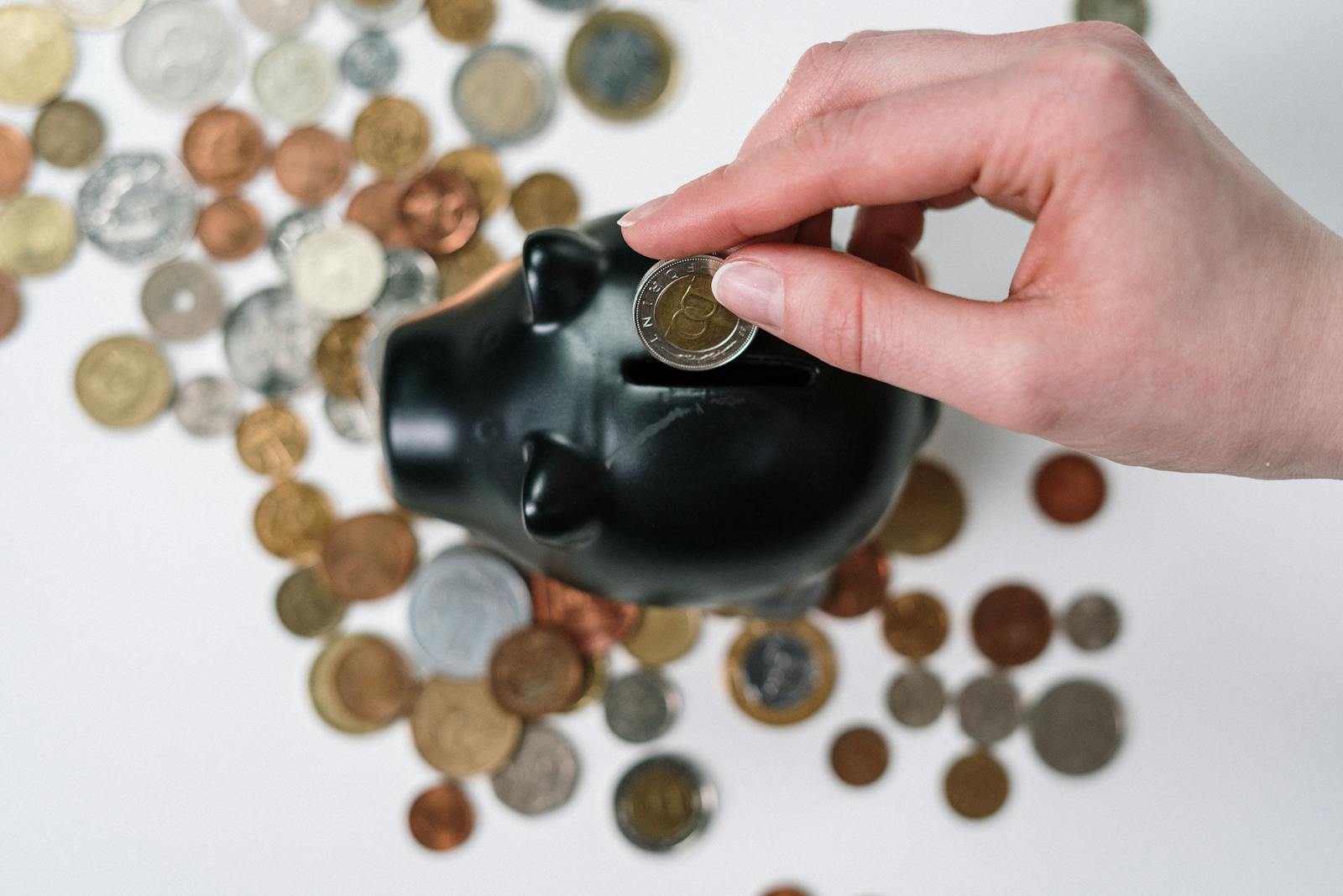 Hand inserting coin into black piggy bank with scattered coins