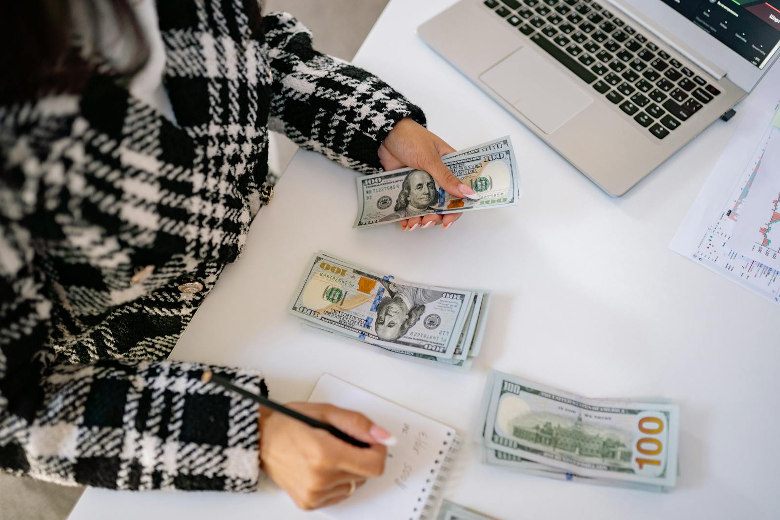 Woman in plaid coat counting American dollars at desk with notebook and laptop