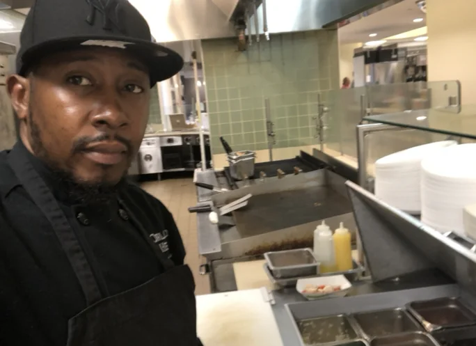 About Dwayne Lindsay 1 Dwayne Lindsay in a black chef's jacket and NY cap, standing beside the flat-top grill and prep line of a commercial kitchen